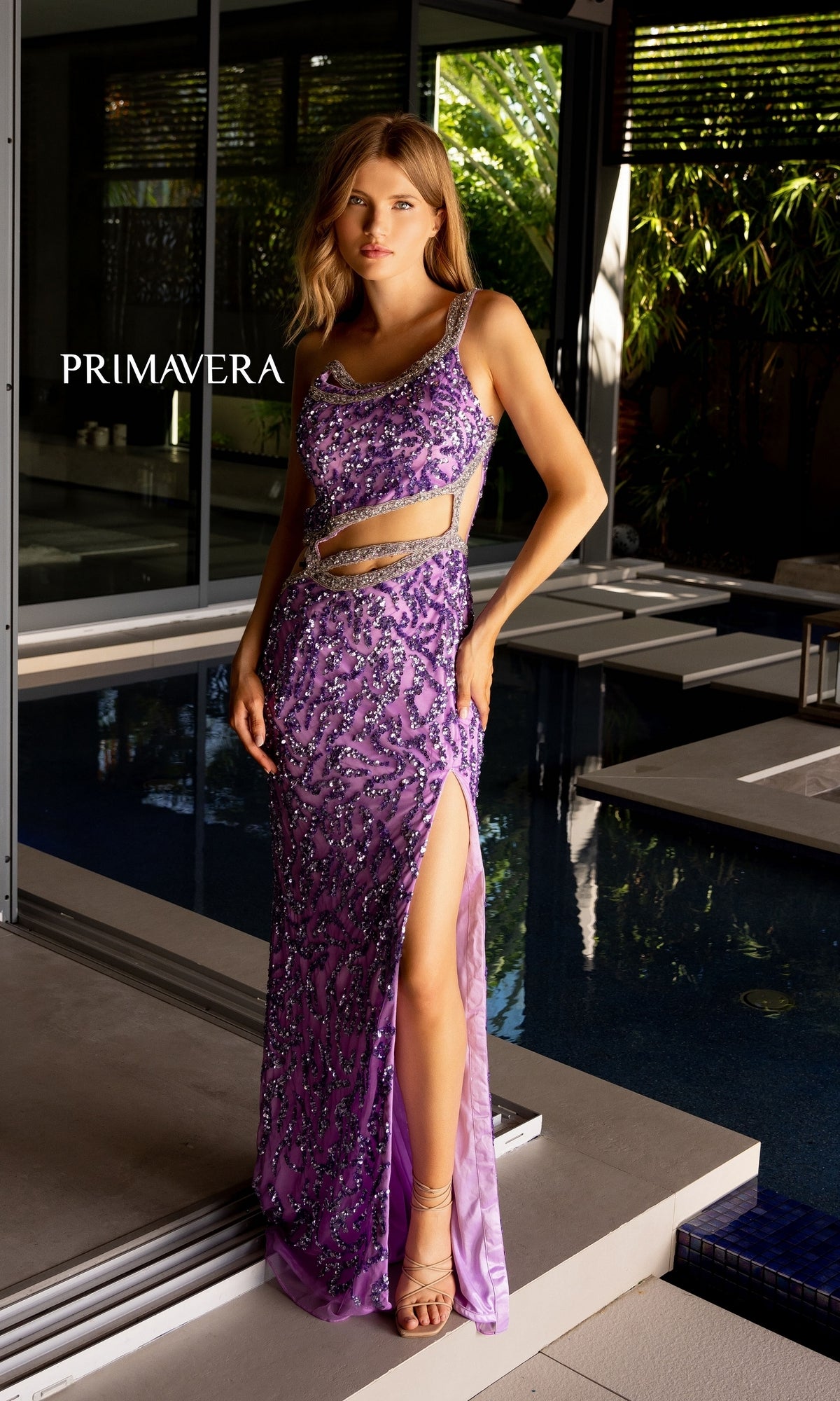 A woman stands gracefully by a modern poolside in the Primavera One-Shoulder Beaded Long Prom Dress 4126, featuring a sleeveless purple design with beaded details, cutout accents, and a side slit.